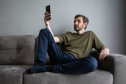 Young man relaxing on a comfortable sofa in his modern living room, holding a mobile phone and looking at the screen, engaging with digital content and enjoying casual leisure time