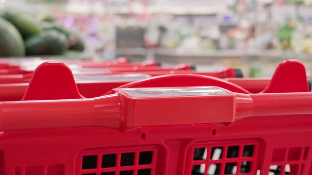 Close-up of a long row of nested red shopping carts with blurred figures of customers walking in the background inside a large modern supermarket or hypermarket. Retail trade and consumerism concept.