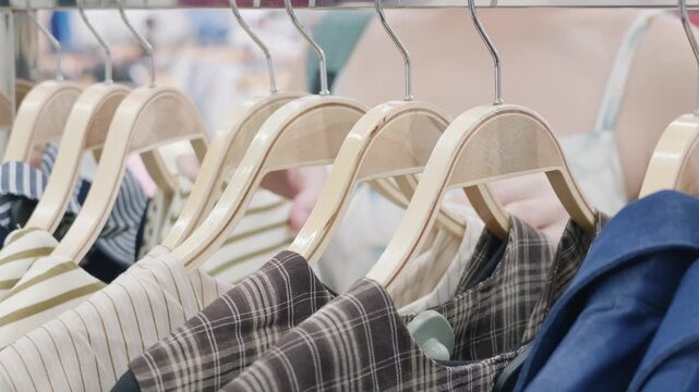 Close-up of wooden hangers with clothes on a rack as a woman browses and chooses dresses in a fashion boutique or clothing store. Retail trade, consumerism and apparel shopping concept.