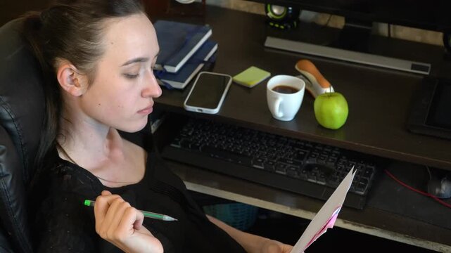 A person is focused on writing notes while sitting at a desk. There is a coffee cup, an apple, and a phone placed nearby. The office has a computer and books around.