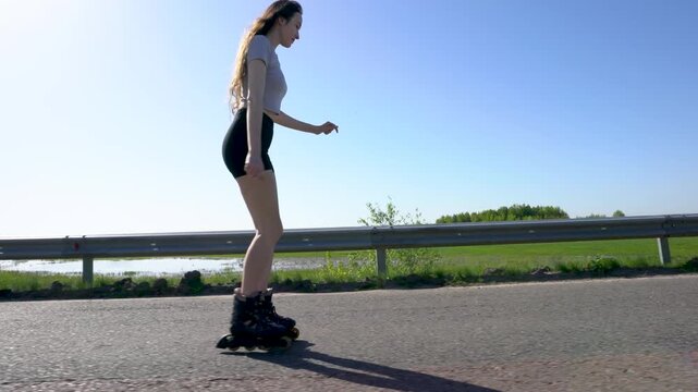 A girl skates on rollerblades along a road under clear blue skies. Green fields and a water body are visible in the background during daylight.