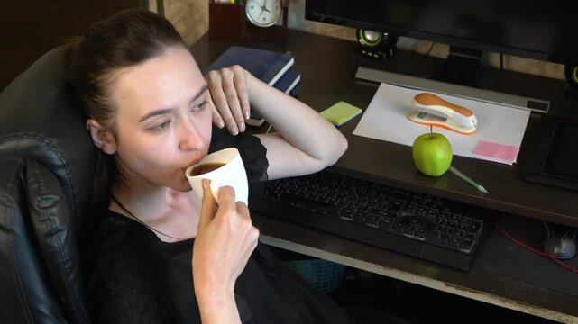 A girl sits at a table in a home office, holds a cup of coffee and looks thoughtful. There is an apple next to the computer, and papers are scattered around.