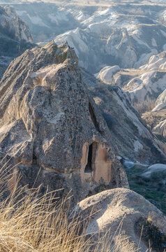 Ancient rock-cut cave dwelling carved into a volcanic tuff formation overlooking a rugged valley in Cappadocia, Turkey, surrounded by distinctive eroded rock formations and sparse winter vegetation.