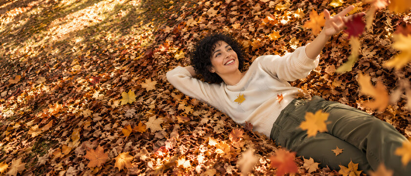 A joyful curly haired woman lies on her back in a carpet of golden autumn leaves, raising one arm to toss colorful foliage into the air while smiling in warm sunlight beneath the trees around her