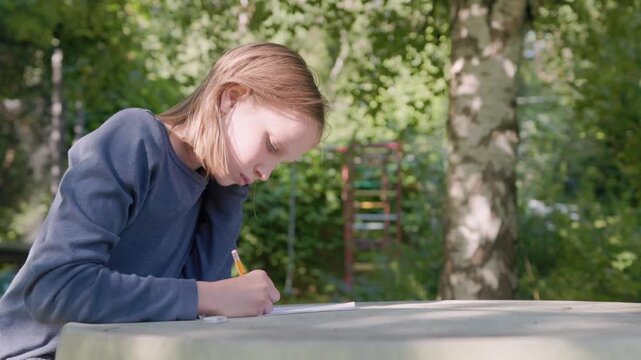 Young girl writes on notepad outdoors at table, showing signs of learning difficulty and deep concentration. Schoolgirl works on homework assignment under bright natural sunlight in park.