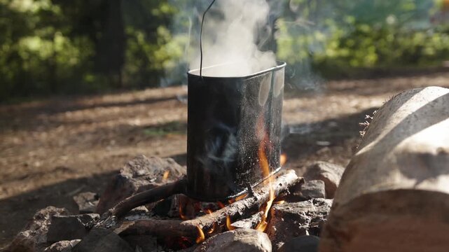 Metal cooking pot heats over campfire flames during outdoor camping trip. Hot water vessel emits thick steam as burning wood warms metal container inside tranquil forest campsite setting.