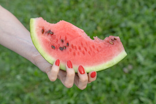 Woman's hand with bright red nails holding a juicy bitten watermelon slice against a natural green background, evoking summer refreshment, healthy snack and outdoor leisure