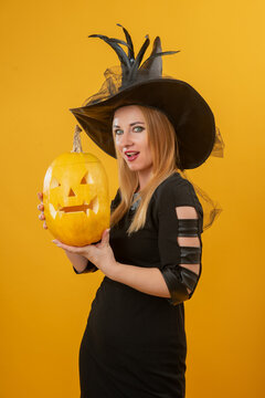 Young blonde woman dressed as a witch, wearing a black hat and dress, holding a carved pumpkin with a spooky face, celebrating the Halloween season against a vibrant orange background