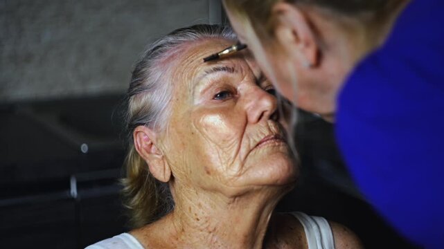Young woman helping an elderly 87 year old woman groom her eyebrows with mascara at home in a kitchen studio highlighting intergenerational care and personal grooming