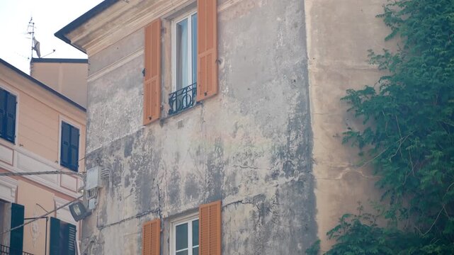 Old masonry building facade stands among neighboring homes while camera pans upward. Open wooden shutters, narrow windows and leafy greenery reveal worn plaster and classic architectural details.