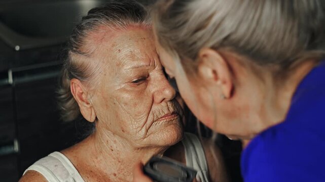 Elderly woman receiving facial skincare and makeup as a young woman applies cream with a brush in a domestic kitchen studio
