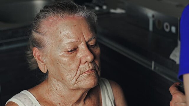 Facial beauty care for an elderly woman as a younger woman carefully applies foundation cream with a brush in a home kitchen setting