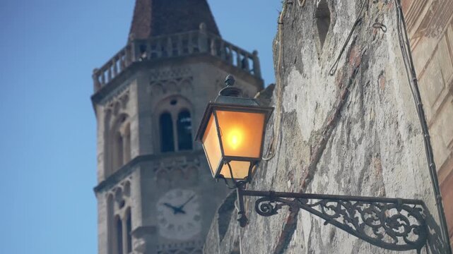 Warm street lantern glows on weathered stone facade near tall clock tower. Decorative metal bracket and arched tower windows lend this quiet architectural view timeless old world character.
