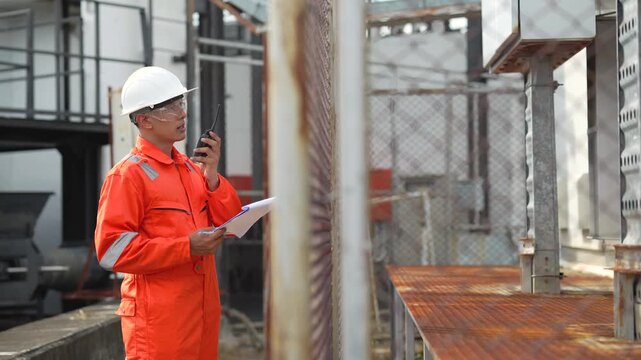 Engineer holds handheld radio and clipboard while inspecting fenced transformer installation at industrial facility. Electrical infrastructure monitoring, maintenance communication, safety inspection.