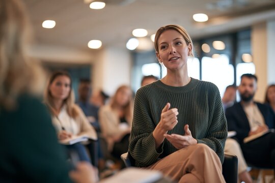 Woman manager speaking to group of employees during mental health first aid workshop in modern office training room with recessed ceiling lights and large windows letting