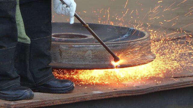 A worker wearing protective clothing and gloves uses an oxy-fuel gas torch to cut a heavy steel train wheel, spraying glowing sparks across an industrial scrap yard.