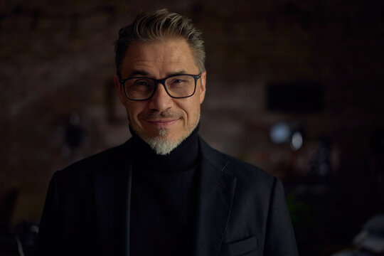 Portrait of happy mid adult man at home in dark room. smiling, Mid adult, middle aged man in 50s smiling, wearing glasses.