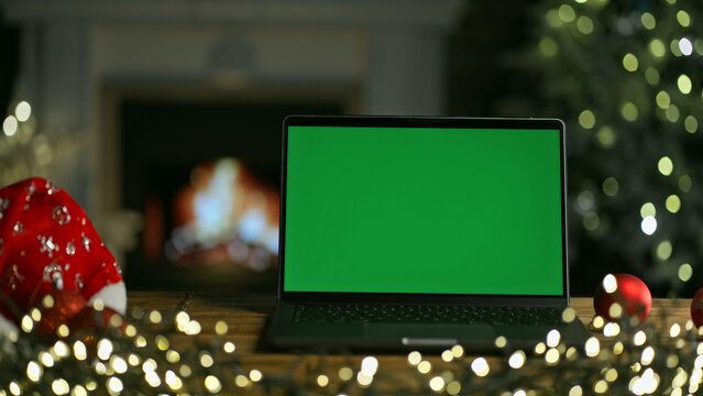 Laptop computer on desk at Christmas. Decorated room with Christmas tree and lights. Cosy winter home with fireplace. Blank green screen for web shop offer and holiday advertisement.