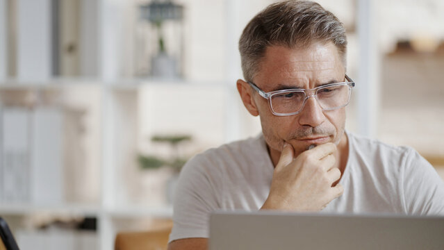Casual mid adult man with laptop computer in home office, banking online, remote working. Portrait of older bearded guy thinking. Businessman, entrepreneur managing business on internet.