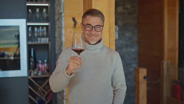 Happy middle aged man in glasses raising a glass of red wine towards camera, smiling in a wooden cabin home. Mature man toasting, celebration, cheers, wine tasting, warm cozy interior.