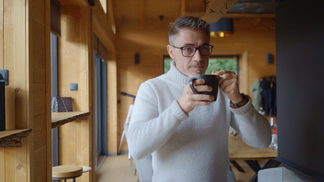Mature man in glasses and white turtleneck sweater, drinking hot coffee or tea in stylish, modern wooden cabin. Senior male enjoying relax time at home. Businessman on mountain vacation, lifestyle.