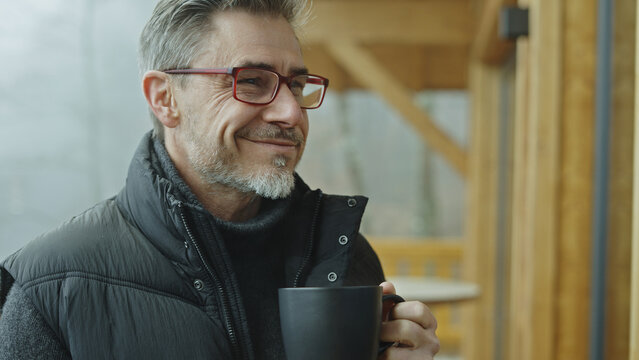 Portrait of middle aged man with gray hair and glasses outdoor on home balcony. Mid adult male drinking hot tea or coffee for getting warm in cold weather. Autumn or winter. Happy satisfied smiling.