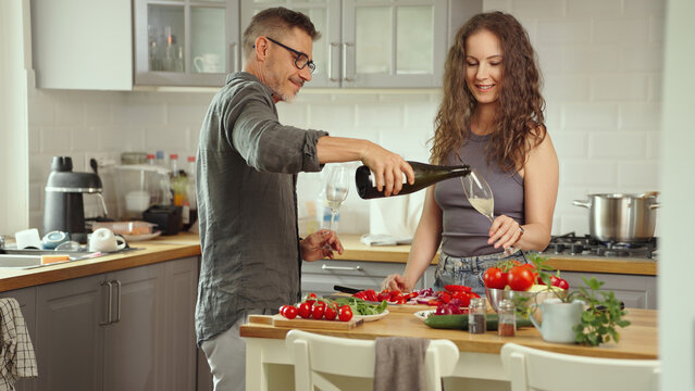 Mid adult couple preparing for cooking in modern kitchen at home, wearing casual cloth. Man and woman following food recipe, cleaning and slicing vegetables, drinking champagne.