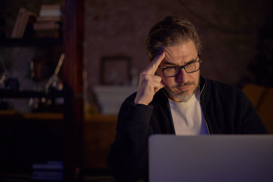 Serious middle aged man with glasses focused on laptop, working late at night. Mature bearded man thinking in cozy living room, face lit by screen. Concentration, home office, remote work.