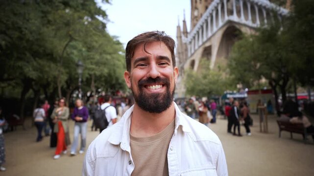 A Caucasian man with a beard smiles at the camera outside Sagrada Familia in Barcelona, Spain. The basilica and visitors are visible behind him.