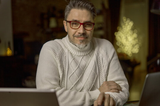 Portrait of mature age, middle age, mid adult casual man in 50s, confident happy smiling, glasses, gray hair. Dark cosy room at home, winter, Christmas.