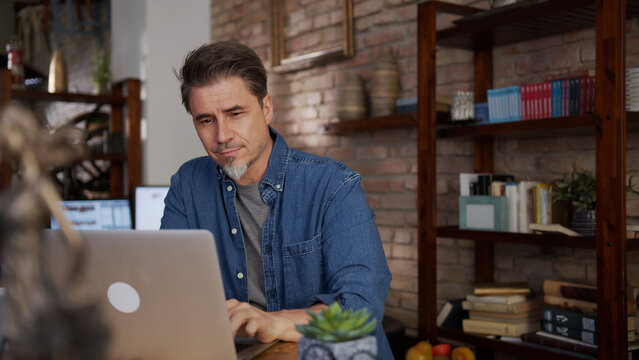 Mature adult man in 50s working on laptop in home office, working from home online, using internet, thinking.