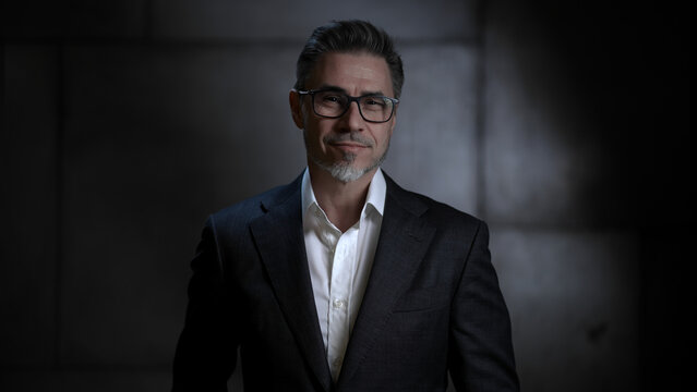 Portrait of happy mature businessman, smiling. Entrepreneur standing in front of dark loft wall. Older, middle aged, mid adult, man in his 40s or 50s in jacket, business casual.