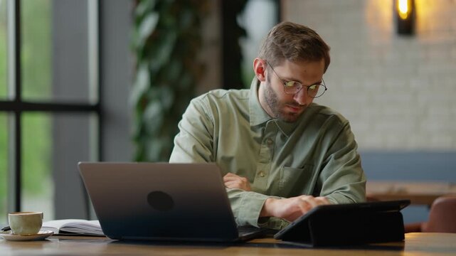 Focused young man working diligently on his laptop in a cozy indoor environment, taking notes and enjoying a warm beverage, showcasing a productive atmosphere.