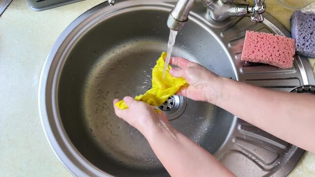 Female hands washing yellow cleaning cloth in kitchen sink. Hand rinsing microfiber cloth under running water stream in metal sink. Housewife performs housework for hygiene at home with cloth.