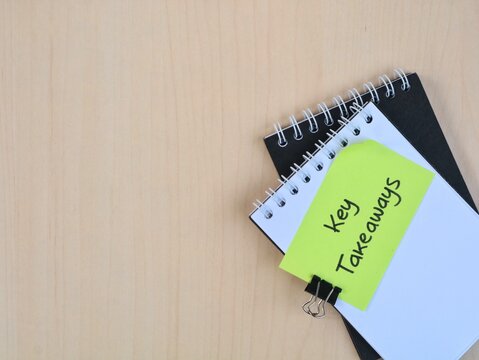 Key takeaways handwritten message on a green adhesive note attached to books on a wooden table, top view. Business summary, learning points, and education concept with copy space