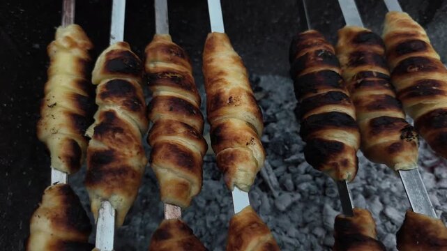 Delicious pastry bakes on metal skewers over hot glowing charcoal embers during outdoor picnic. Golden dough roll browns evenly under open fire heat. Fresh pastry provides tasty snack treat