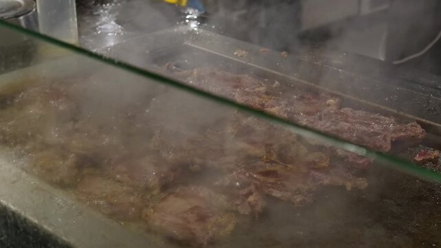 Close up view of multiple pieces of raw beef steak sizzling and cooking on hot commercial metal griddle surface with visible steam rising in professional restaurant kitchen environment setting.