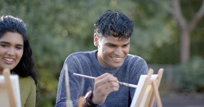 Indian couple painting on tabletop easels in park, man starting strokes, woman mirroring, bonding