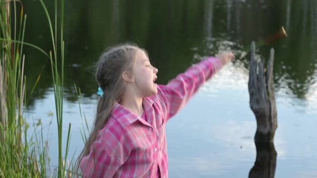 Young girl imagines magical sorcerer spells with cattail plant at river bank. Little kid wearing pink shirt holds organic reed stalk outside. Small youth explores fantasy imagination in countryside.