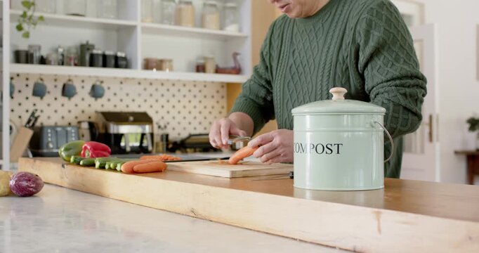 Mature Asian man peeling carrots at home camera pullback adding peels to compost bin for cooking