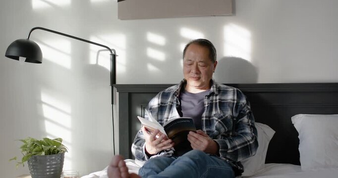Middle-aged Asian man in flannel shirt on bed reading paperback, glancing at sunlight pattern