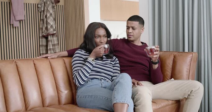 African American couple on sofa sharing, woman offering mug after sipping, man checking and tasting