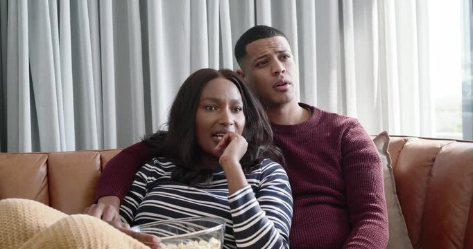 African American couple sitting on sofa reacting to TV, pointing and sharing clear bowl of popcorn