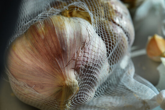 Macro shot focusing on the texture of a garlic bulb inside a white plastic mesh bag with soft natural light. High detailed culinary background.