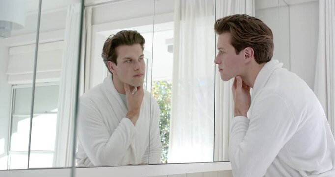 Adult man in white robe noticing self in 3-panel mirror, raising chin and palpating neck, grooming