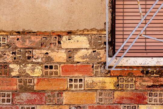 An exterior building wall displays exposed red and yellow perforated clay bricks joined by rough gray mortar below a smooth beige plaster section. On the right side a window features closed horizontal