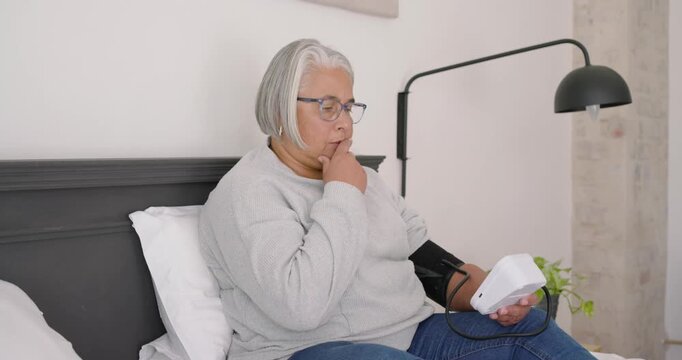 Senior African American woman sitting on bed, seeing BP monitor, entering data on phone, confirming