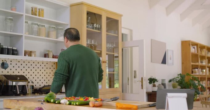 Asian man in green sweater chopping on board at island, following tablet instructions, pouring oil