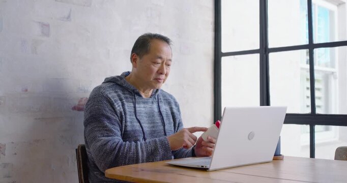During remote call, Asian man holding white bottle, red cap, confirming label on laptop at table
