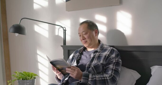 Asian man sitting on bed by window reading paperback while sunlight shifting and causing quiet gaze